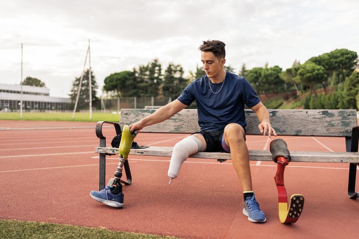 Stock photo of disabled man athlete sitting in a bench and putting on his leg prosthesis.